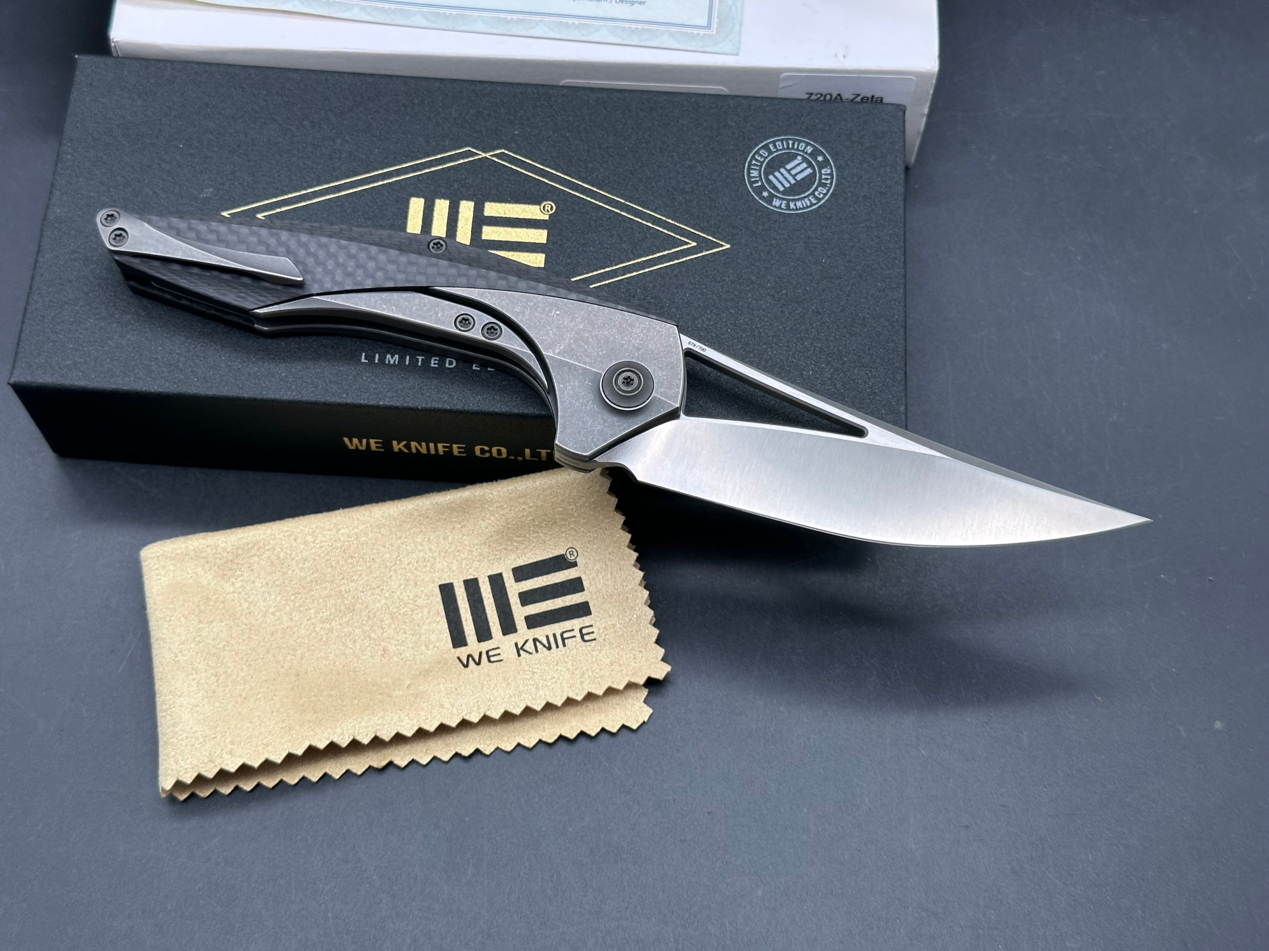We Zeta: Elijah Isham design. 3.875 inch M390, trailing point, flat grind, satin finish blade. Frame lock. Carbon fiber and titanium handle
